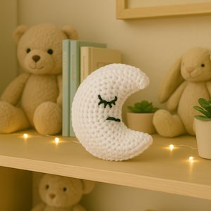 Könnte beinhalten: Ein weißer, gehäkelter Halbmond mit schwarzen, gestickten Augen und Mund, der auf einem Holzregal liegt. Der Mond ist ein dekorativer Gegenstand, möglicherweise für ein Kinderzimmer, zusammen mit Stofftieren und Büchern.