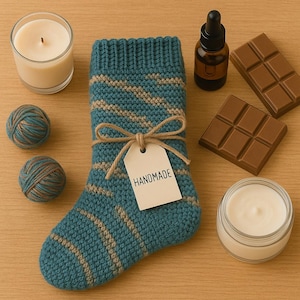 May include: A teal and tan knitted stocking tied with twine and a tag that reads "HANDMADE". The image also includes a candle, two balls of yarn, a bottle of oil, two chocolate bars, and a jar of cream, all arranged on a wooden surface.