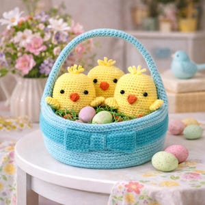 Könnte beinhalten: Ein gehäkelter hellblauer Osterkorb mit drei gelben Küken mit orangefarbenen Schnäbeln und schwarzen Augen. Der Korb ist mit grünem Kunstgras und pastellfarbenen Eiern gefüllt. Ein Schleifendetail ziert die Vorderseite des Korbs.