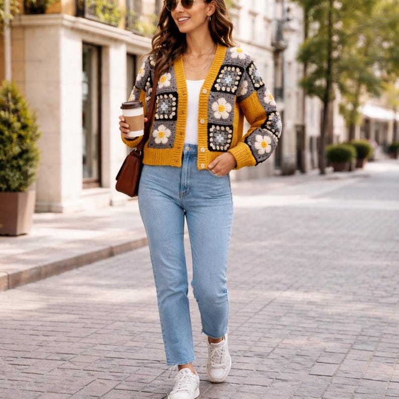 May include: A mustard yellow cardigan with a floral granny square design. The cardigan has long sleeves and a button-up front. The model is wearing light blue jeans and white sneakers. A brown purse is over the shoulder.