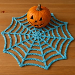 Könnte beinhalten: Ein geschnitzter orangefarbener Kürbis mit Stiel liegt auf einem hellblauen gehäkelten Spinnennetz. Das Spinnennetz besteht aus dickem Garn und wird auf einer Holzoberfläche präsentiert. Der Kürbis hat dreieckige Augen und einen geschnitzten Mund.
