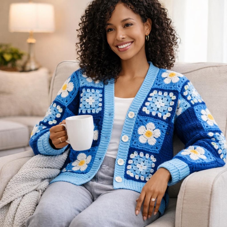 May include: A blue crochet cardigan with white daisy flower squares. The cardigan has a light blue trim and buttons. The person in the image is holding a white mug. The cardigan is a cozy and stylish garment.