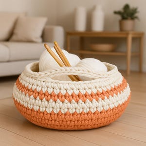 Puede incluir: Una cesta de ganchillo hecha a mano en crema y naranja, llena de ovillos de lana blancos y agujas de tejer de madera. La cesta tiene un asa y un patrón geométrico. Perfecta para guardar materiales de manualidades o como elemento decorativo.