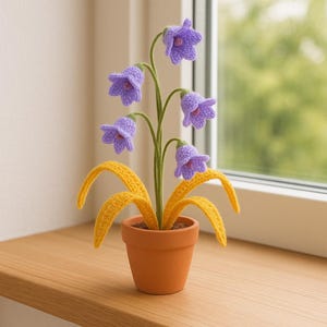 Può includere: Una composizione floreale all'uncinetto fatta a mano con fiori viola a forma di campana, foglie gialle e uno stelo verde, in un piccolo vaso di terracotta. La composizione è su una superficie di legno vicino a una finestra.