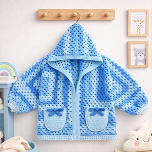 May include: A blue and white crochet hooded jacket with two pockets, each adorned with a bow. The jacket is hanging on a wooden rack. The crochet pattern is a granny square design. The jacket is a light blue color.