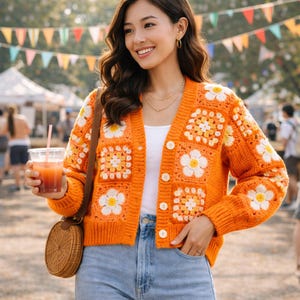 May include: Orange crochet cardigan with white daisy flower squares. The open-front sweater has a V-neck and is buttoned. The woman is wearing a white top and blue jeans. She is holding a drink and a brown woven purse.