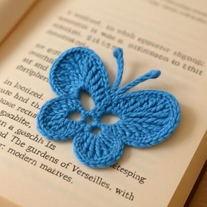 Może przedstawiać: Ręcznie robiona, niebieska zakładka do książki w kształcie motyla, leżąca na otwartych stronach książki. Motyl ma szczegółowe wzory skrzydeł i czułki. Strony książki są postarzane, z widocznym tekstem.