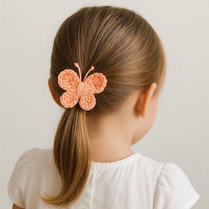 May include: A peach-colored, crocheted butterfly hair clip is attached to a ponytail. The butterfly has detailed wings and antennae. The child is wearing a white shirt. The background is a plain, light color.
