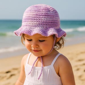 Könnte beinhalten: Ein helllila gehäkelter Sonnenhut mit gerüschtem Rand und Kinnbändern. Der Hut wird von einer Person getragen, die ein weißes Tanktop an einem Strand trägt. Der Hut ist aus weichem, strukturiertem Garn gefertigt.