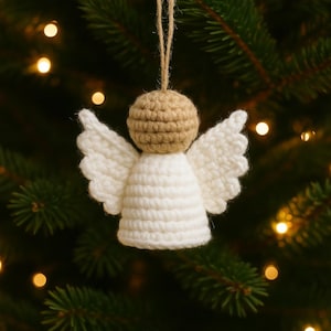 May include: A handmade crochet angel Christmas ornament. The angel has a tan head, white wings, and a white body. It is suspended by a natural twine string. The background is a blurred Christmas tree with soft, glowing lights.