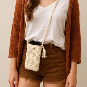 Może przedstawiać: Kremowy, szydełkowy pokrowiec na telefon z pasującym paskiem i frędzlami. Etui posiada zapięcie na guzik i jest przeznaczone do noszenia na szyi lub na ramieniu. Pokrowiec na telefon jest pokazany na białej koszuli i brązowych szortach.