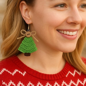 May include: Green crochet Christmas tree earrings with a brown trunk and a wooden button detail. The earrings are suspended from gold-colored hooks and adorned with a twine bow. The earrings are a festive accessory.