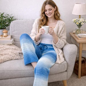 May include: A woman wearing blue and white ombre crochet pants and a cream-colored cardigan, holding a white mug. The pants have a drawstring waist. She is sitting on a gray armchair.