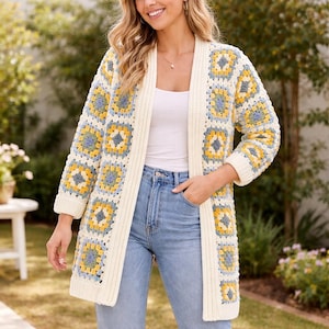 Könnte beinhalten: Lange, offene Häkeljacke mit Granny-Square-Muster. Die Strickjacke hat eine cremefarbene Basis mit gelben und blauen Quadraten. Die Ärmel und Kanten sind cremefarben. Die Strickjacke wird über einem weißen Oberteil und einer blauen Jeans getragen.