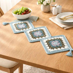 May include: A handmade crocheted table runner in a square pattern, featuring blue, white, and brown hues, with tassels at each end. It's laid on a wooden table, accompanied by a bowl of grapes, plates, and cutlery.