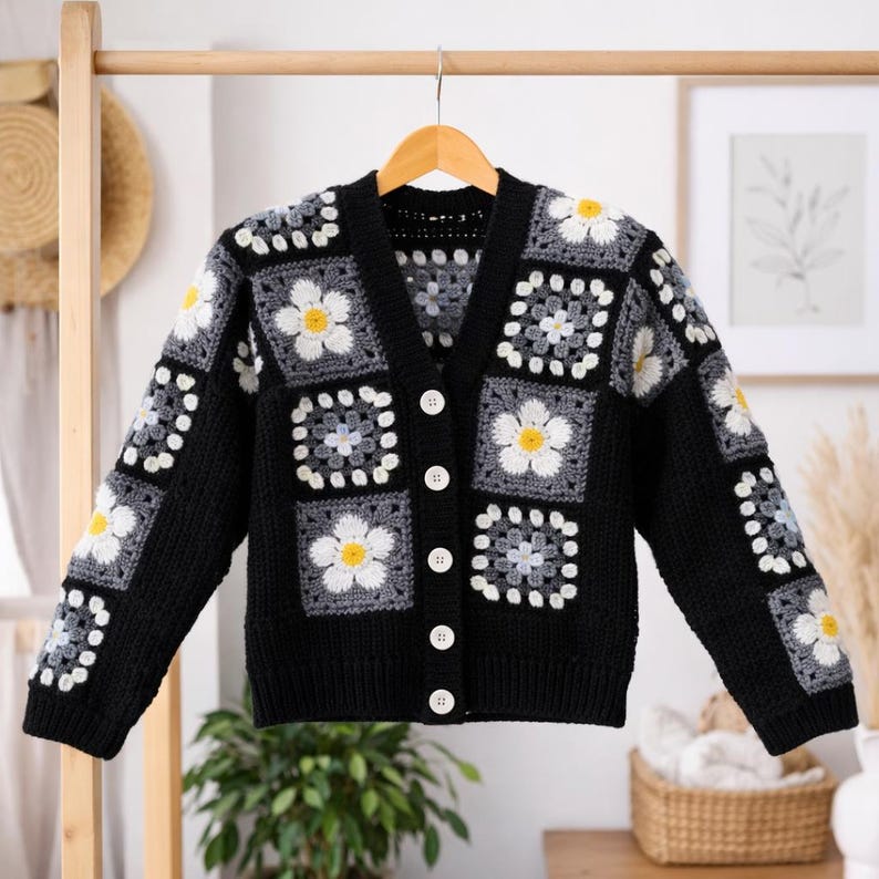 May include: A black crochet cardigan with a daisy pattern. The cardigan features white daisies with yellow centers on gray and white squares. It has a V-neck, long sleeves, and white buttons down the front. The garment is hanging on a wooden hanger.