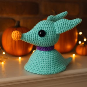May include: A crocheted teal-colored dog-like figure with a purple collar and orange nose. The figure has large black eyes and pointed ears. Two orange pumpkins are in the background, along with small, warm-toned lights.