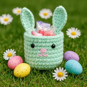 May include: A mint green crochet Easter basket shaped like a bunny, filled with candy. The basket has black eyes and a pink nose. Colorful speckled Easter eggs and daisies are scattered around the basket on a bed of green grass.