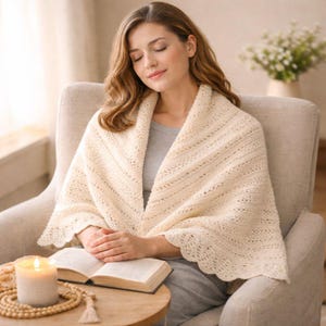 Puede incluir: Un chal de punto color crema con borde festoneado está drapeado sobre los hombros. El chal lo lleva una persona sentada en un sillón beige. Una vela encendida, un libro abierto y un collar de cuentas descansan sobre una pequeña mesa de madera.