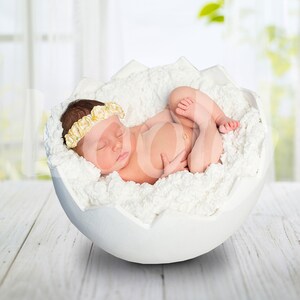 May include: A newborn baby wearing a yellow flower headband sleeps peacefully in a white egg-shaped bed.