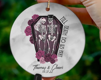 May include: A round white ceramic ornament featuring a graphic of two skeletons in a coffin with dark pink roses. The text reads Till Death Do Us Part, Thomas & Cheri, and 10.37.22. A gold string is attached to the top for hanging purposes.