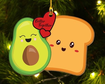 May include: A flat hanging ornament featuring a cartoon avocado and a slice of toast side by side. Both have smiling faces and rosy cheeks. A red heart shape is positioned between them with the black text Better Together printed on it.