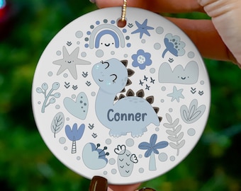 May include: A round white ceramic ornament featuring a light blue cartoon dinosaur with the name Conner printed on its side. The design includes various blue and grey motifs such as a rainbow, stars, flowers, clouds, and leaves, with a gold cord.