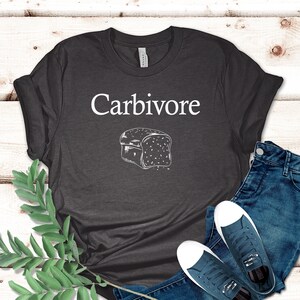 May include: A dark gray t-shirt with the word "Carnivore" printed in white on the front. A white outline of a loaf of bread is below the word. The shirt is laying on a white wooden surface with a pair of blue jeans and a pair of blue sneakers.