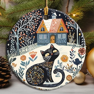Könnte beinhalten: Ein rundes Keramikornament mit einer schwarzen Katze mit komplizierten Gold- und Weißmustern, die in einer verschneiten Landschaft sitzt. Hinter der Katze steht ein Haus mit blauer Fassade und rotem Dach, umgeben von stilisierten Bäumen und Blumen.