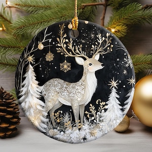 May include: A round Christmas ornament featuring a deer with ornate gold antlers and body details, set against a black background with white trees and snowflakes. The ornament is trimmed with gold and has a gold hanging loop.