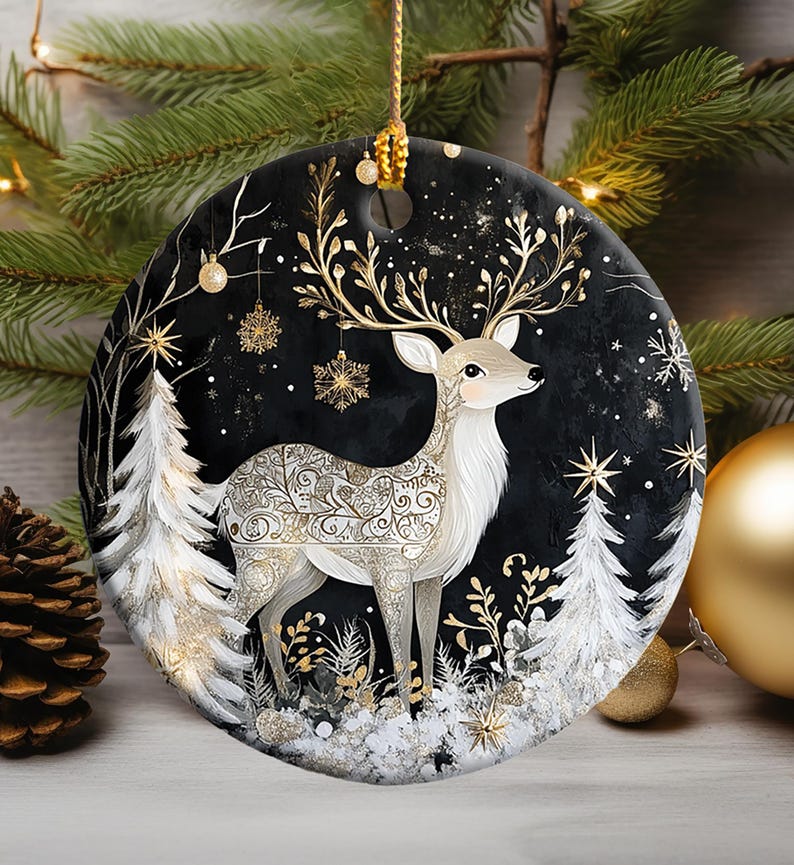 May include: A round Christmas ornament featuring a deer with ornate gold antlers and body details, set against a black background with white trees and snowflakes. The ornament is trimmed with gold and has a gold hanging loop.