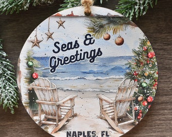 Puede incluir: Un adorno redondo de madera con una ilustración en acuarela de dos sillas de playa frente al océano. El texto dice Seas & Greetings y NAPLES, FL. La escena incluye un árbol de Navidad decorado, estrellas colgantes y adornos.
