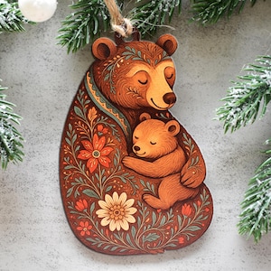 Könnte beinhalten: Ein Holzanhänger in Form eines Bären mit einem Jungtier, mit einer detaillierten Illustration eines großen braunen Bären, der ein kleineres Jungtier umarmt, verziert mit orangefarbenen und gelben Blumen und grünen Blattmustern, an einer braunen Schnur.