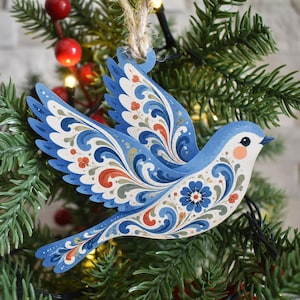 K&ouml;nnte beinhalten: Ein flacher, vogelf&ouml;rmiger Holzanh&auml;nger mit komplexen floralen Rankenmustern in Blau, Wei&szlig; und Orange. Der Vogel ist im Flug mit nach oben ausgebreiteten Fl&uuml;geln dargestellt und an einer Schlaufe aus nat&uuml;rlichem Jutegarn befestigt.