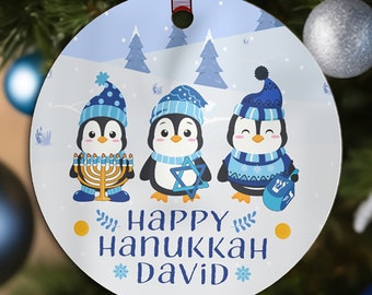 May include: A round, white ornament with a blue winter scene featuring three penguins wearing hats and holding a menorah, a Star of David, and a dreidel. The text "Happy Hanukkah David" is printed on the ornament.