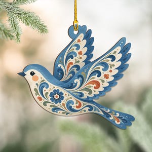 K&ouml;nnte beinhalten: Ein flacher, vogelformiger H&auml;ngeschmuck mit einem floralen Rankenmuster in Blau, Creme und Orange. Der Vogel ist im Flug mit nach oben ausgebreiteten Fl&uuml;geln dargestellt und an einer d&uuml;nnen goldenen Kordel befestigt, die durch ein Loch f&uuml;hrt.