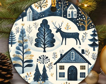 May include: A round ceramic ornament featuring a winter scene with a dark blue deer, evergreen trees, and houses on a cream background. The design includes gold-colored accents on the trees and windows, with a gold string loop at the top for hanging.