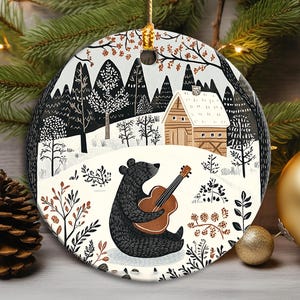 Könnte beinhalten: Ein rundes Keramikornament mit einem schwarzen Bären, der eine braune Akustikgitarre in einer verschneiten Landschaft spielt. Die Illustration zeigt eine Holzhütte, schwarze Tannen und kahle Äste auf weißem Grund, aufgehängt an einer goldenen Kordel.
