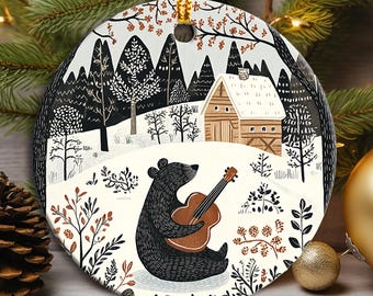 May include: A round ceramic ornament featuring a black bear playing a guitar in a snowy forest scene. The bear is sitting on a snowy patch with a small cabin in the background. The ornament is decorated with a white background and black, brown, and red details.