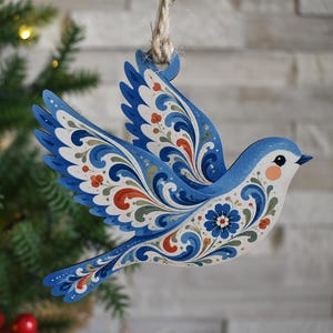 K&ouml;nnte beinhalten: Ein flacher, vogelformiger Holzanh&auml;nger, der an einer Juteschnur h&auml;ngt. Der Vogel ist in Blau-, Wei&szlig;- und Oranget&ouml;nen bemalt und mit komplizierten Blumen- und Wirbelmustern auf den Fl&uuml;geln und dem K&ouml;rper verziert, inklusive einer kleinen blauen Blume.