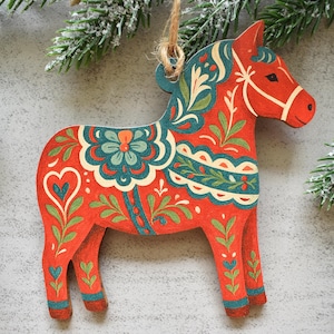 Pode incluir: Um ornamento de madeira em forma de cavalo Dala, pintado num vermelho vibrante com padrões florais intrincados em azul-petróleo, creme e verde. O cavalo apresenta uma crina estilizada, um motivo de coração na garupa e um laço de juta.