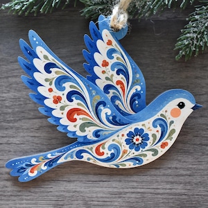 K&ouml;nnte beinhalten: Ein flacher, vogelf&ouml;rmiger H&auml;ngeschmuck mit komplexen floralen Mustern und Schn&ouml;rkeln in Blau, Wei&szlig; und Orange. Der Vogel ist im Flug mit ausgebreiteten Fl&uuml;geln dargestellt und verf&uuml;gt &uuml;ber eine kleine Schlaufe aus Jutekordel zum Aufh&auml;ngen.