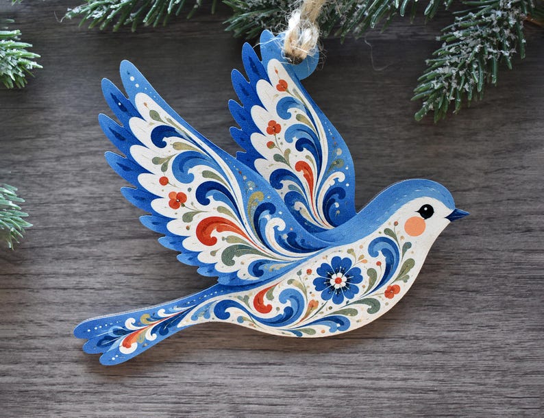 K&ouml;nnte beinhalten: Ein flacher, vogelf&ouml;rmiger H&auml;ngeschmuck mit komplexen floralen Mustern und Schn&ouml;rkeln in Blau, Wei&szlig; und Orange. Der Vogel ist im Flug mit ausgebreiteten Fl&uuml;geln dargestellt und verf&uuml;gt &uuml;ber eine kleine Schlaufe aus Jutekordel zum Aufh&auml;ngen.