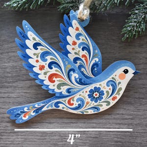K&ouml;nnte beinhalten: Ein flaches, vogelformiges Ornament in Blau mit komplexen floralen Schn&ouml;rkelmustern in Wei&szlig;, Orange und Gr&uuml;n. Das Ornament hat eine Schlaufe aus Jutekordel zum Aufh&auml;ngen und wird mit einer 10,16 cm Messlinie am unteren Bildrand gezeigt.