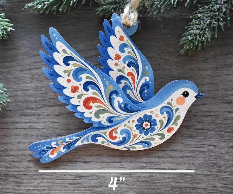 K&ouml;nnte beinhalten: Ein flaches, vogelformiges Ornament in Blau mit komplexen floralen Schn&ouml;rkelmustern in Wei&szlig;, Orange und Gr&uuml;n. Das Ornament hat eine Schlaufe aus Jutekordel zum Aufh&auml;ngen und wird mit einer 10,16 cm Messlinie am unteren Bildrand gezeigt.
