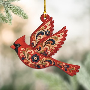 May include: A flat, red cardinal bird ornament hanging from a gold string. The bird is depicted in flight with wings spread, featuring intricate, swirling floral patterns in shades of dark blue, cream, and muted green across its body and wings.