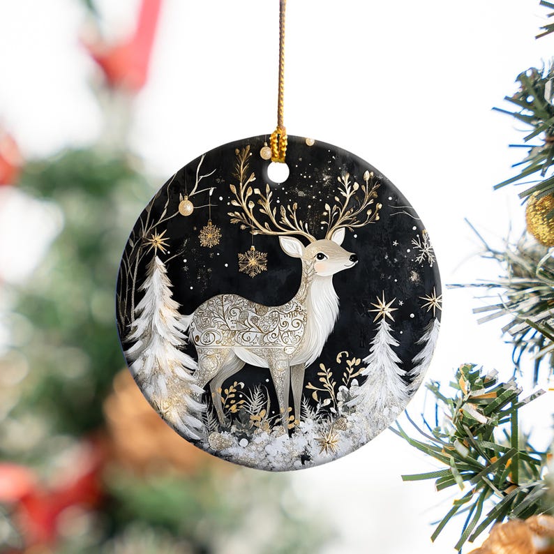 May include: A round Christmas ornament featuring a detailed illustration of a deer with ornate gold antlers and body patterns. The deer stands in a snowy forest scene with white trees and gold accents against a black background. A gold string is attached for hanging.