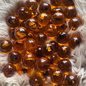 May include: A collection of amber-colored glass marbles, some with embedded insect designs. The marbles vary in transparency, with some appearing darker than others. The marbles are scattered on a fluffy, light-colored surface.