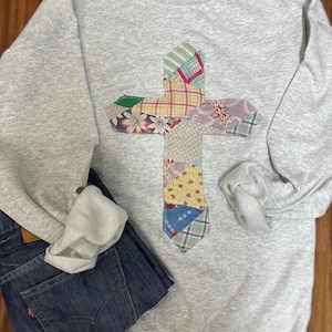 May include: A light gray crewneck sweatshirt featuring a large cross applique made from various colorful fabric patches with floral, plaid, and geometric patterns. The sweatshirt is paired with dark blue denim jeans with a red brand tag.