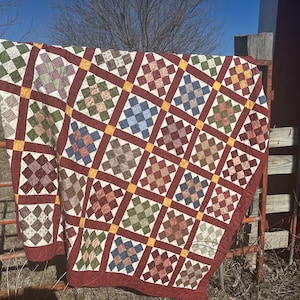 May include: A quilt with a patchwork design featuring squares of various colors and patterns, including red, green, blue, and brown. The quilt is hanging on a rusty metal fence.
